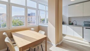 Una cuina moderna amb armaris blancs, una taula de menjador de fusta i vista a edificis a través de grans finestres.