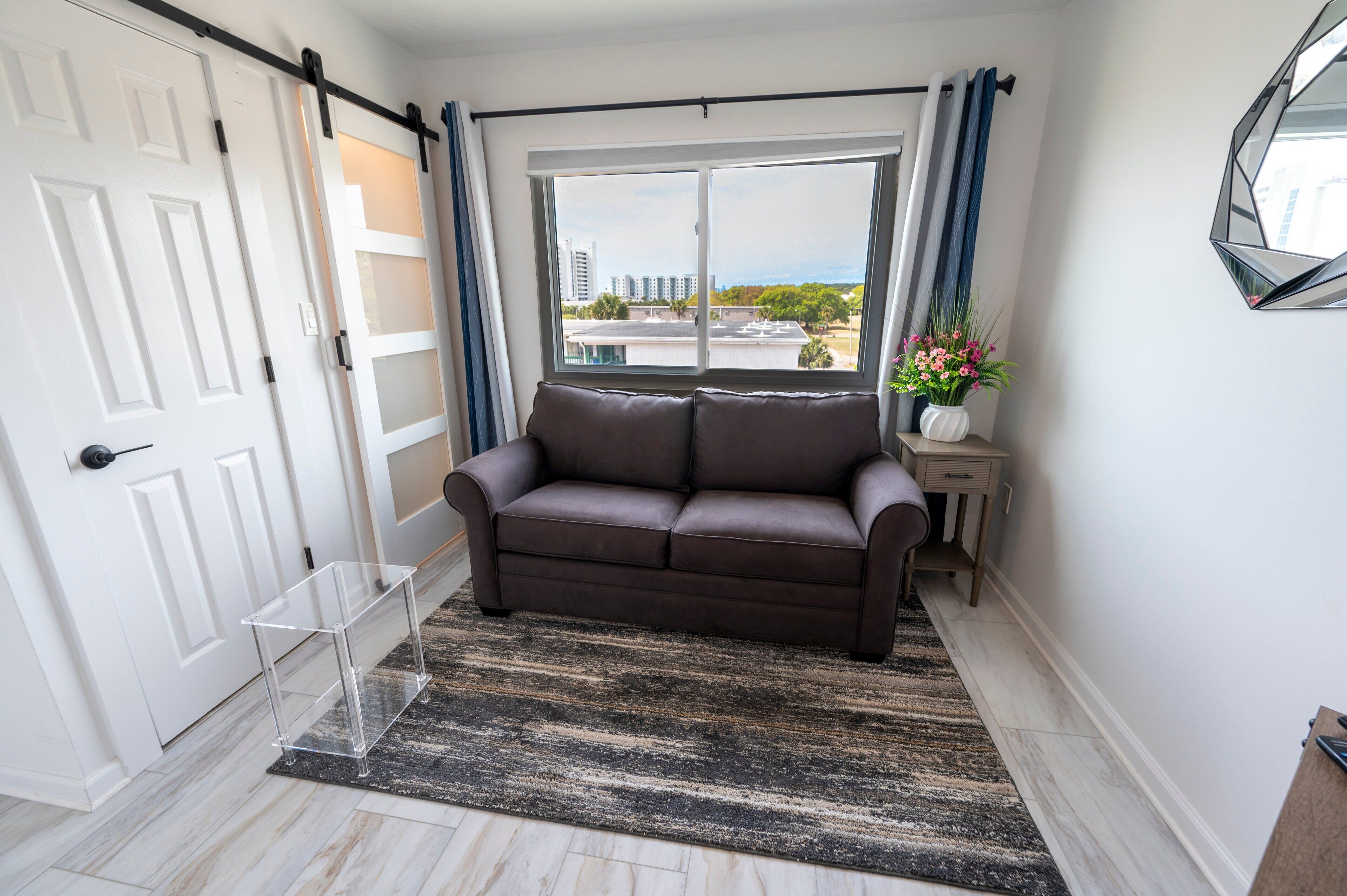 A room with a brown sofa, a clear glass table, a large window with curtains, and a view of buildings outside.