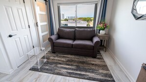 Una habitación con un sofá marrón, una mesa de vidrio transparente, un ventanal con cortinas y vistas a edificios.