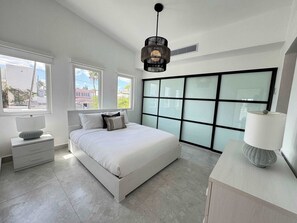Ein modernes Schlafzimmer mit einem großen Bett, zwei Nachttischen, einem Beistelltisch, einem großen Fenster und einem Deckenventilator.