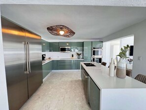 Eine moderne Küche mit einem Edelstahlkühlschrank, einer Mikrowelle und einem Backofen. Weiße Arbeitsplatte mit Spüle und einer Vase mit Blumen.