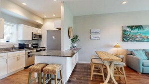 A modern kitchen with white cabinets, a refrigerator, and a microwave. There is a wooden dining table with stools and a painting of a beach scene on the wall. - Shared Pools, Boardwalk to the Beach, Golf Cart Access (Port Aransas)