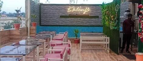An outdoor seating area with tables and chairs, a neon sign, and decorative plants.