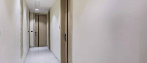 Ένας στενός, καλά φωτισμένος διάδρομος με απλούς τοίχους και πόρτες, που οδηγεί σε ένα δωμάτιο.