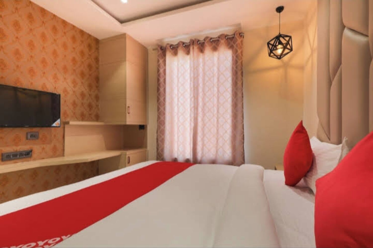 A hotel room with a bed, a TV mounted on the wall, a window with curtains, and a hanging light fixture.