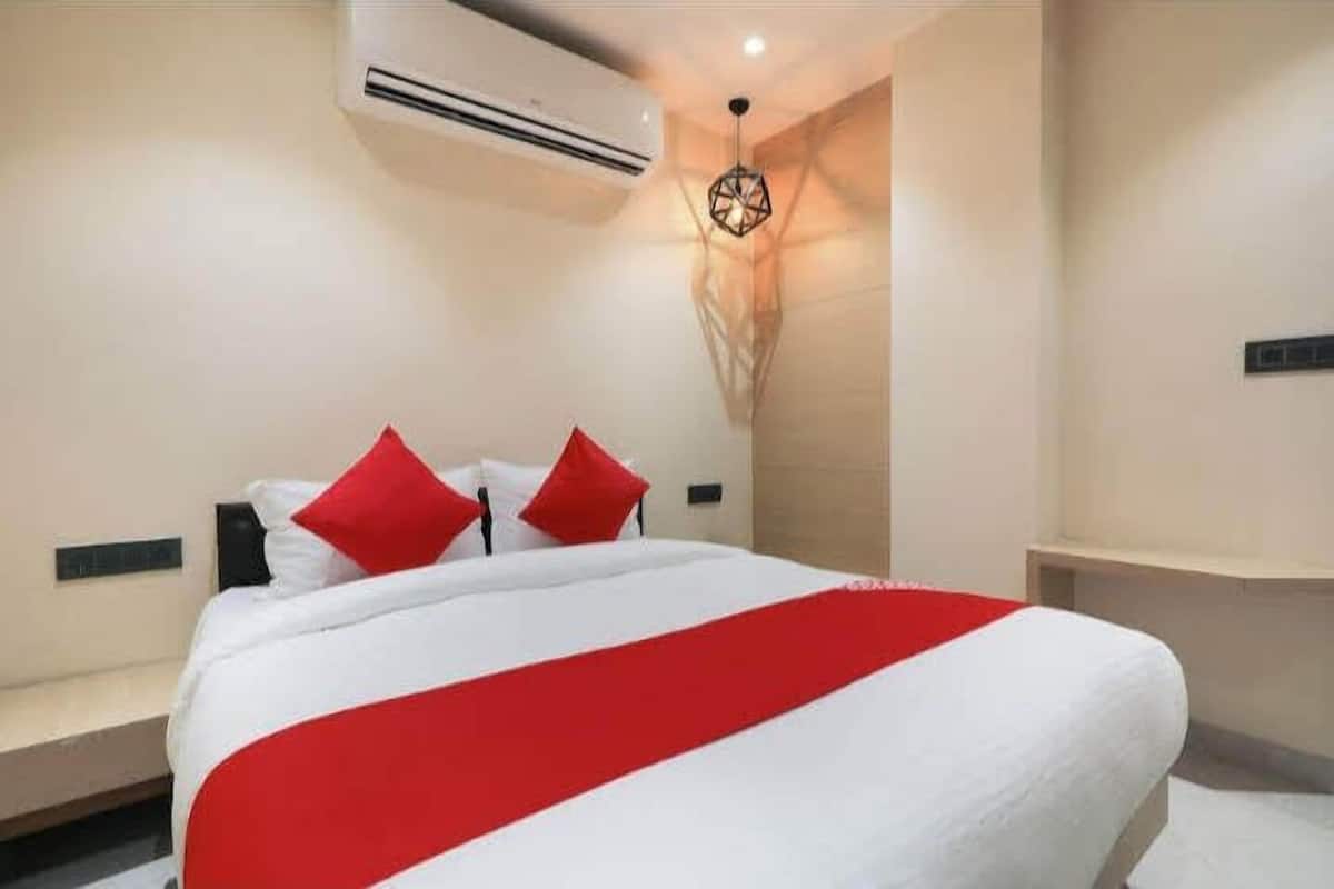 A hotel room with a bed, red pillows, a white bedspread, an air conditioning unit, and a unique wall-mounted light fixture.