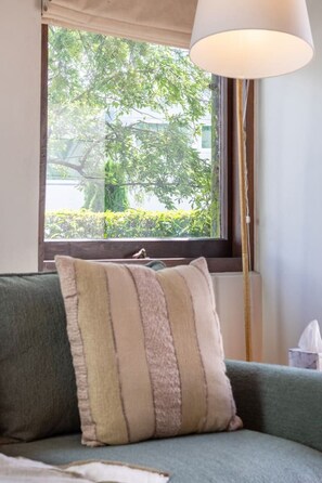 Ein Zimmer mit einem grünen Sofa, einem beige-braunen Streifenpolster, einer Stehlampe und einem Fenster mit Blick auf Grün.