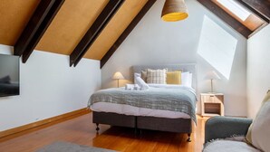 A bedroom with a sloped ceiling, a bed with a grey and white duvet, two bedside lamps, and a TV mounted on the wall. - Merivale | King Bed + Carpark, Walk to Hagley Park (Christchurch)