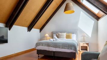 A bedroom with a sloped ceiling, a bed with a grey and white duvet, two bedside lamps, and a TV mounted on the wall.