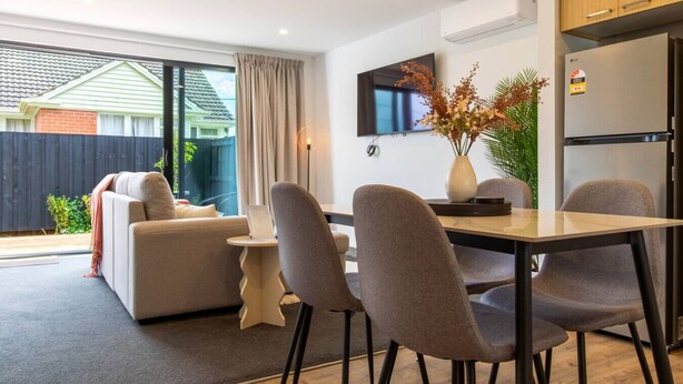 A modern living room with a sofa, dining table, and chairs. There is a TV mounted on the wall, a vase with flowers on the table, and a refrigerator in the kitchen area. - 2-Bedroom Townhouse Opposite The Palms + Carpark (Christchurch)