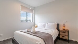 A modern bedroom with a large bed, a bedside table with a lamp, and a window with a view of the city. - 2-Bedroom Townhouse Opposite The Palms + Carpark (Christchurch)