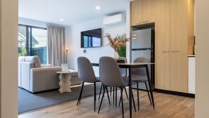 A modern living room with a sofa, a dining table with chairs, a flat-screen TV mounted on the wall, and a built-in wooden cabinet. - 2-Bedroom Townhouse Opposite The Palms + Carpark (Christchurch)