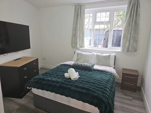 Ein Schlafzimmer mit Bett, Fernseher, Fenster mit Vorhängen und einem kleinen Nachttisch.