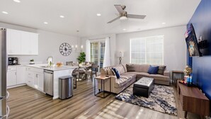 Um apartamento moderno com cozinha, sala de jantar e sala de estar. Possui uma ilha grande na cozinha, uma mesa de jantar com cadeiras, um sofá e uma mesa de centro. Há um ventilador de teto, um relógio e uma janela com persianas.