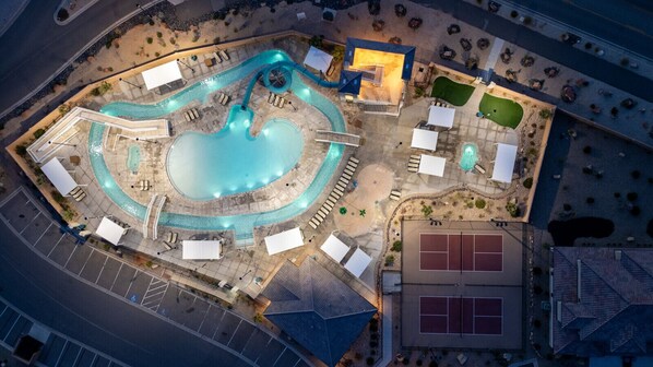 Ein Luftbild eines Resorts mit großem Schwimmbad, Liegestühlen, einem Tennisplatz und einer begrünten Anlage mit Bäumen und Sträuchern.