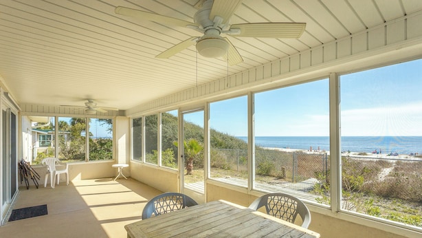 A covered patio with a wooden table and chairs, large windows offering a view of the beach and ocean, and a ceiling fan. - FSU Football, Fall Breezes & Scalloping Adventures (Alligator Point)