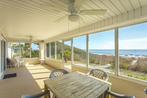 A covered patio with a wooden table and chairs, large windows offering a view of the beach and ocean, and a ceiling fan. - FSU Football, Fall Breezes & Scalloping Adventures (Alligator Point)