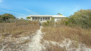 A two-story house with a front porch, surrounded by tall grasses and a sandy path leading to the entrance. - FSU Football, Fall Breezes & Scalloping Adventures (Alligator Point)