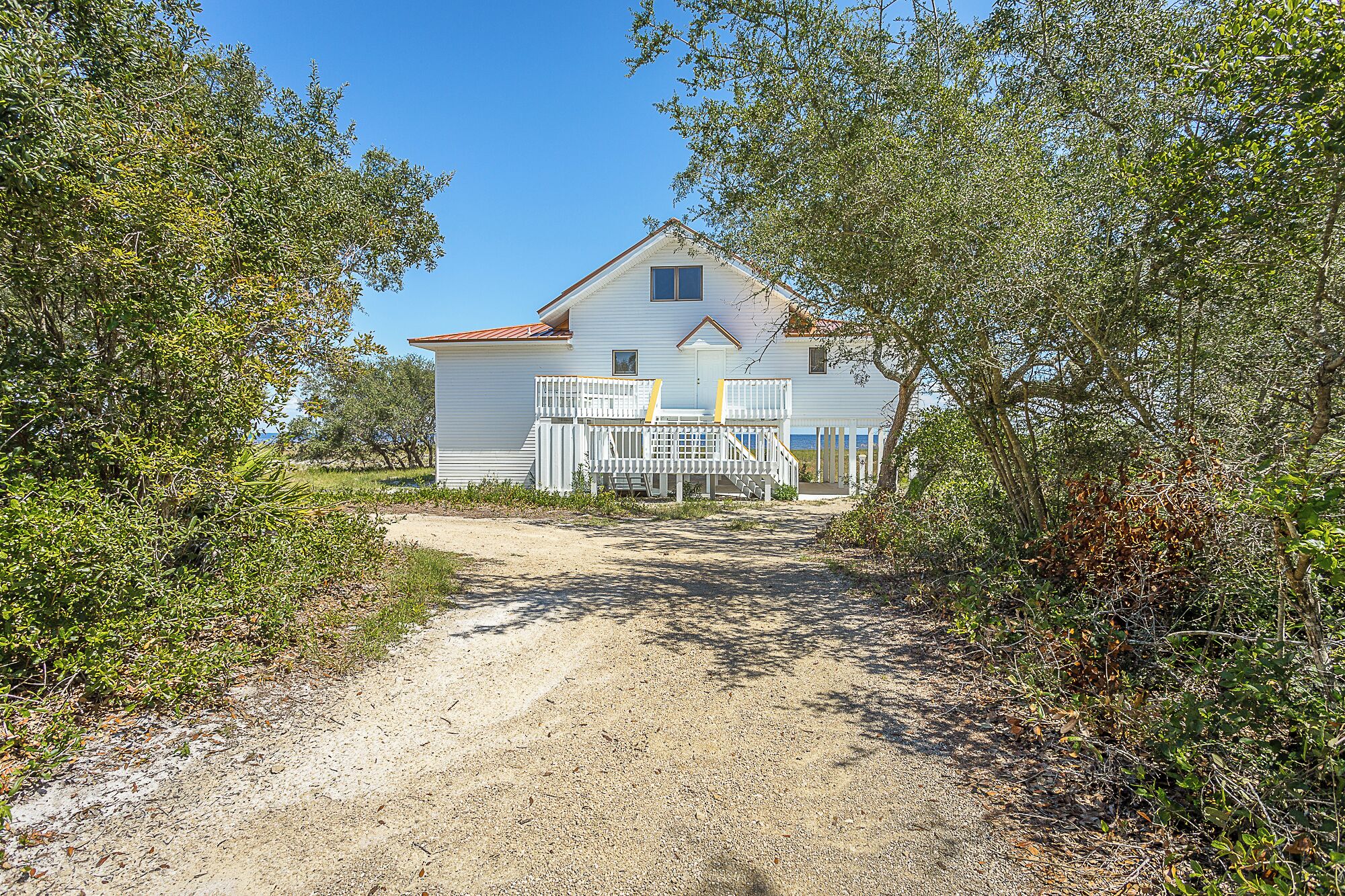 A white house with a porch and a dirt path leading to it, surrounded by trees and shrubs.
