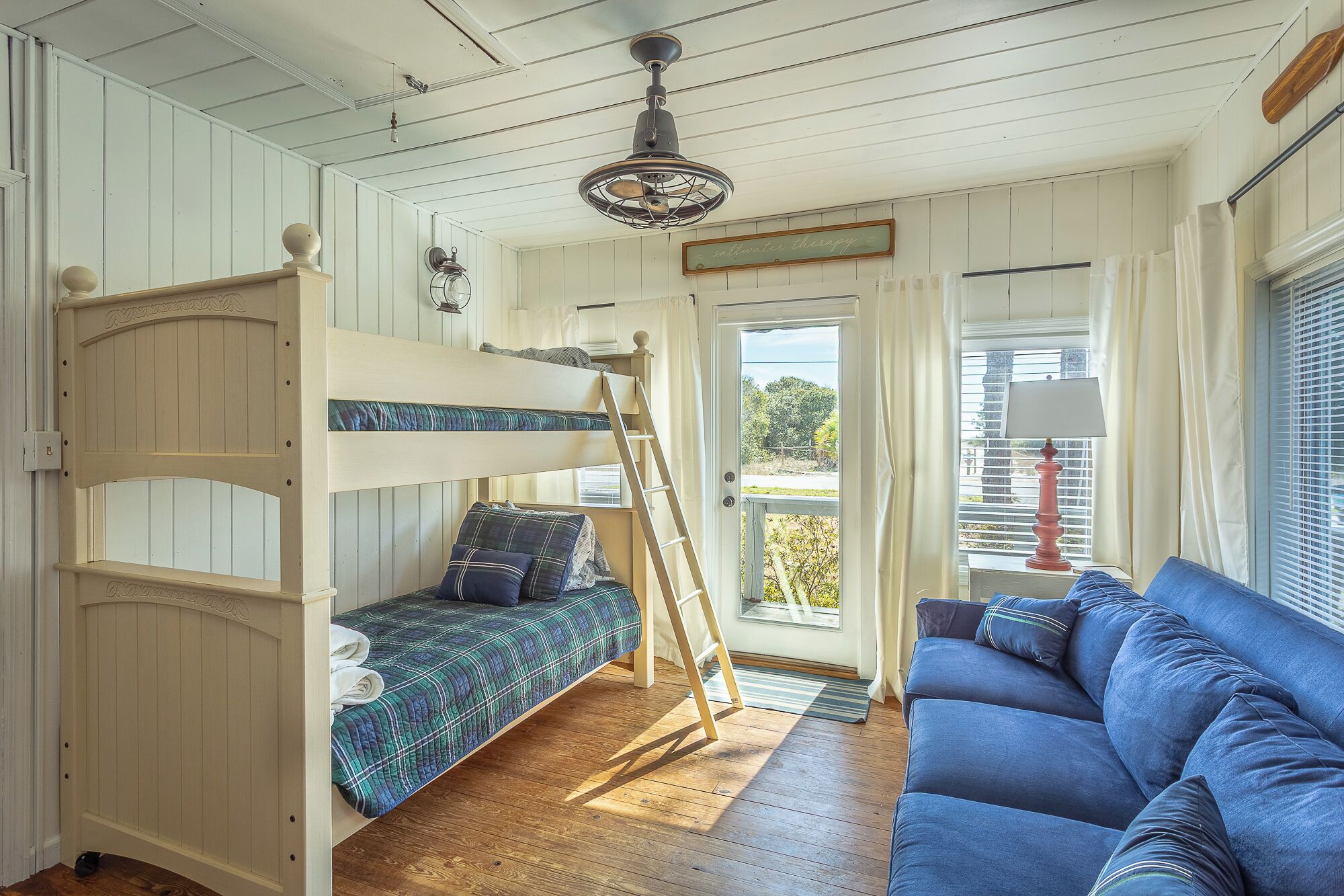 Une pièce avec un lit superposé, un canapé bleu, une échelle en bois, un ventilateur de plafond et une fenêtre avec des rideaux.