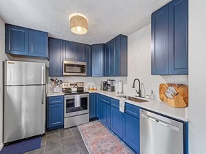 A compact kitchen with blue cabinets, stainless steel appliances, and a small rug. - 5-Star Elegant 4BR: Fresh Reno, Superb View & Clean (Maggie Valley)