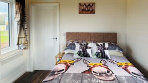 Un dormitorio con techo de madera, una puerta blanca, una ventana con cortinas, una cama con colcha de motivos florales y animales, y un aire acondicionado de pared.