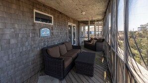 Een overdekt terras met rieten meubels, een houten vloer en grote ramen met uitzicht op het omliggende landschap.