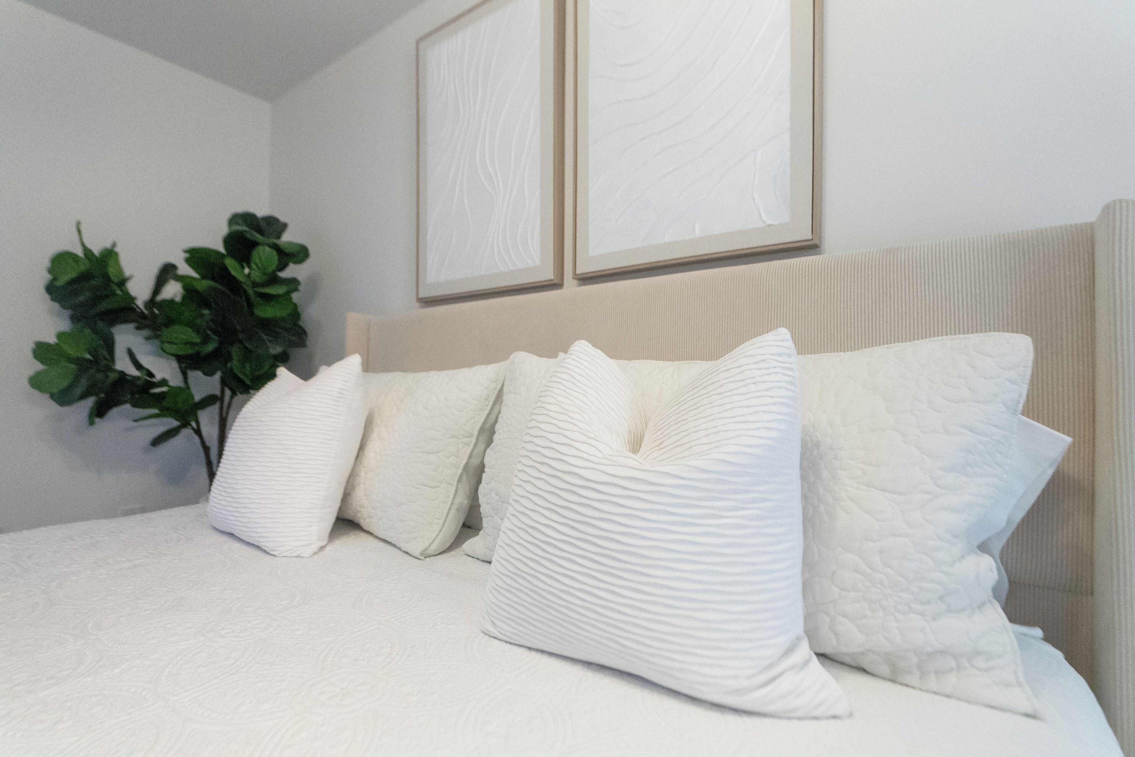 Un lit bien fait, avec une literie blanche et plusieurs oreillers, une plante fixée au mur et deux œuvres d’art encadrées au-dessus du lit.