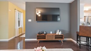 A modern living room with a flat-screen TV mounted on the wall, a wooden entertainment unit below, and a dining area with a chair and table.