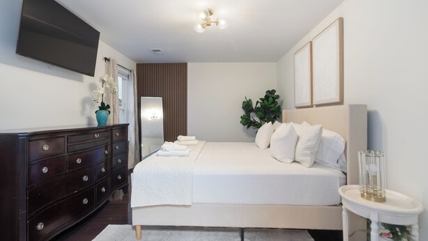A bedroom with a bed, dresser, TV, and decorative plants.