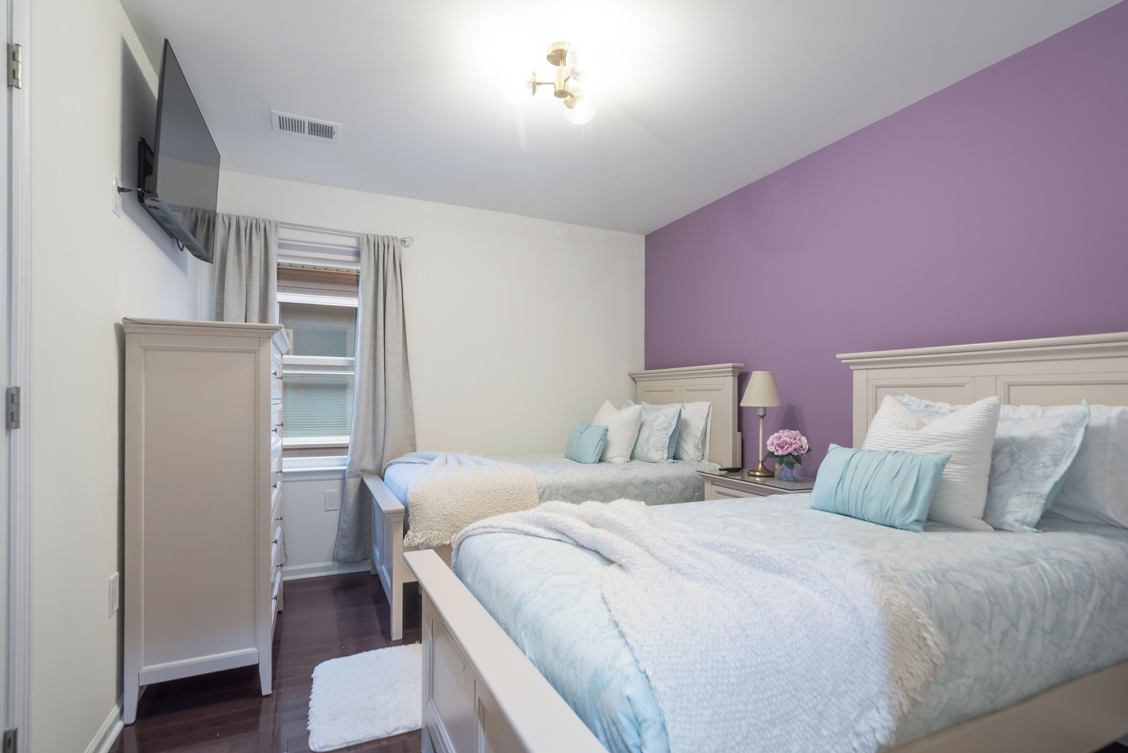 Un dormitorio con dos camas, un armario, un televisor instalado en la pared y una ventana con cortinas.