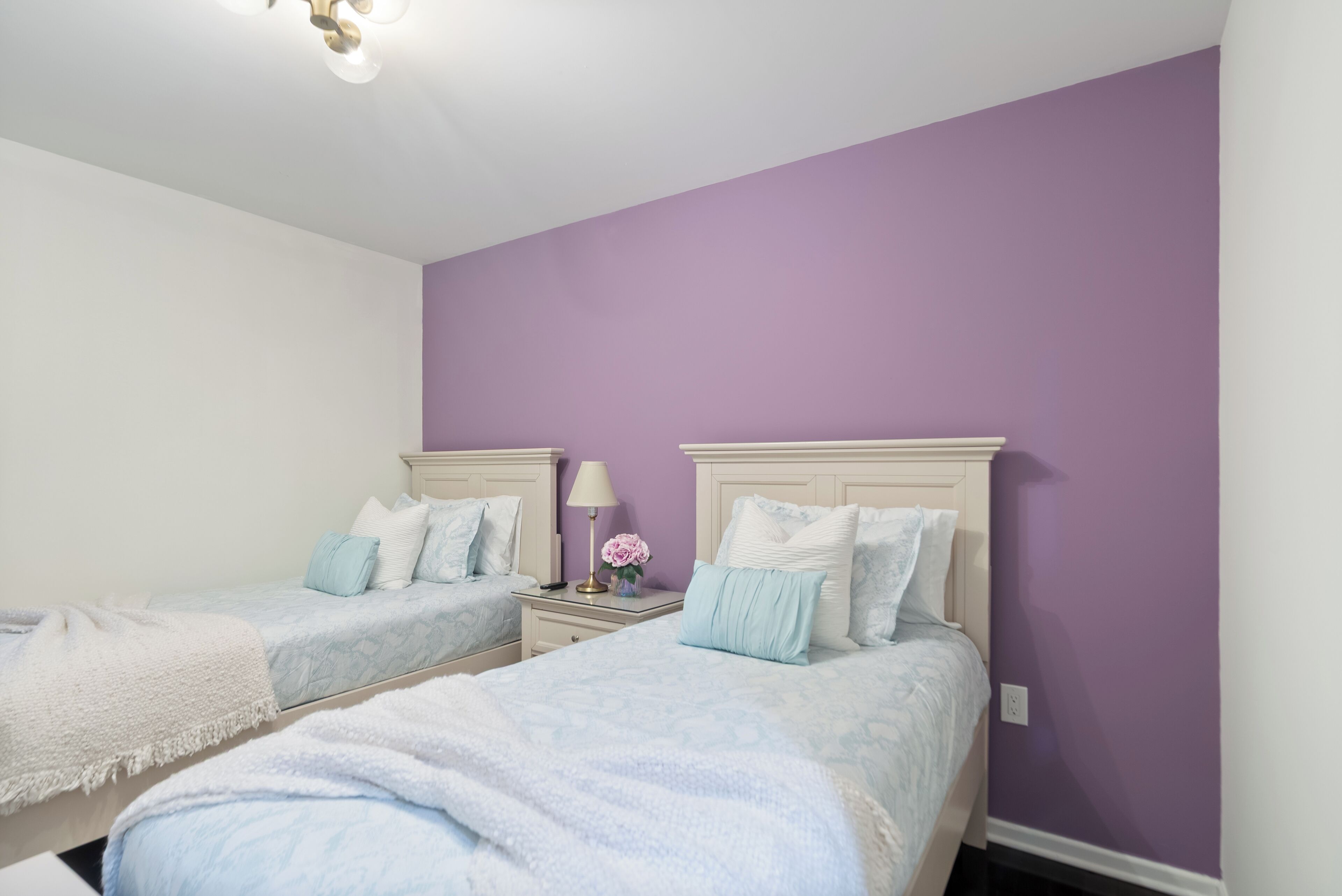 A bedroom with two beds, a nightstand with a lamp and flowers, and a ceiling light fixture.