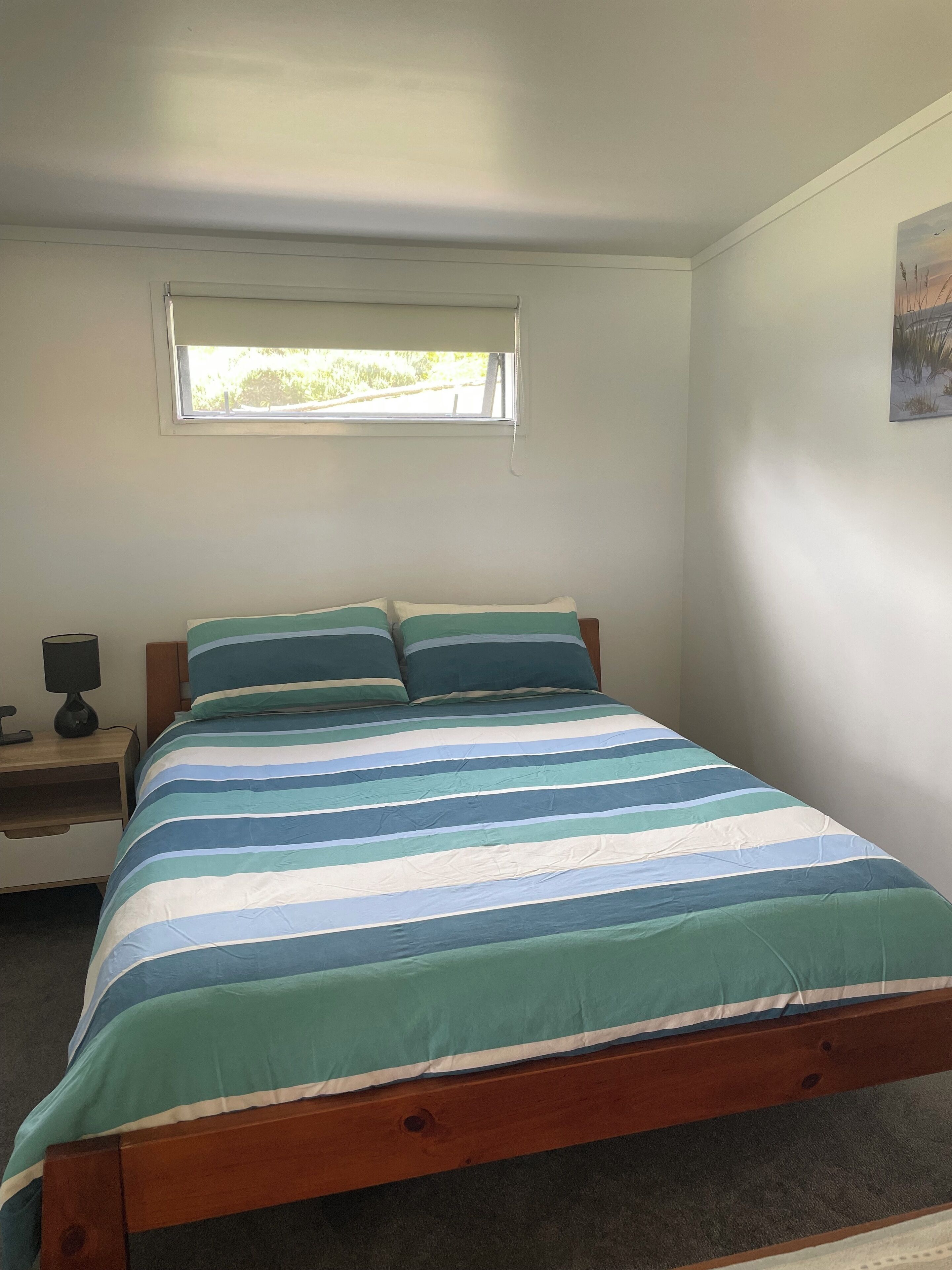 A bedroom with a bed, a nightstand, a lamp, a window, and a painting on the wall.