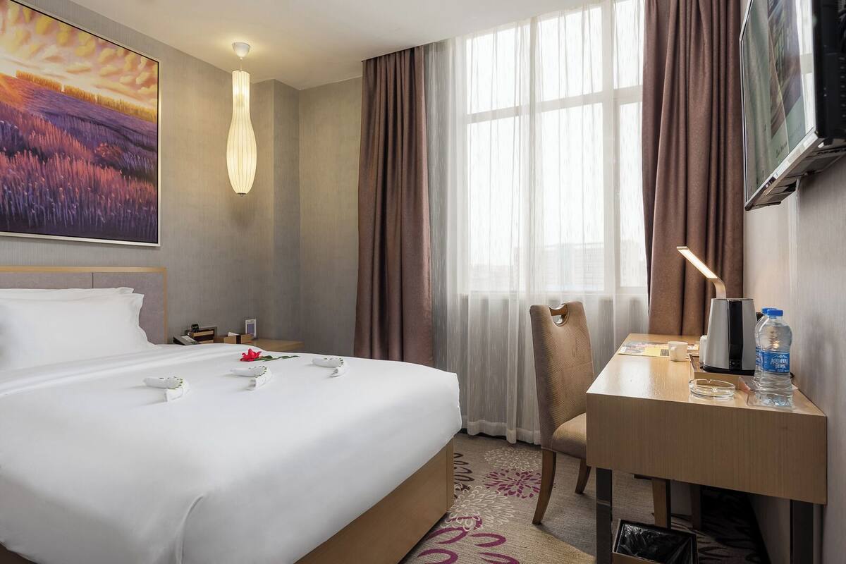 Quarto de hotel com uma cama grande, uma escrivaninha, uma cadeira, televisão e janela com cortinas.