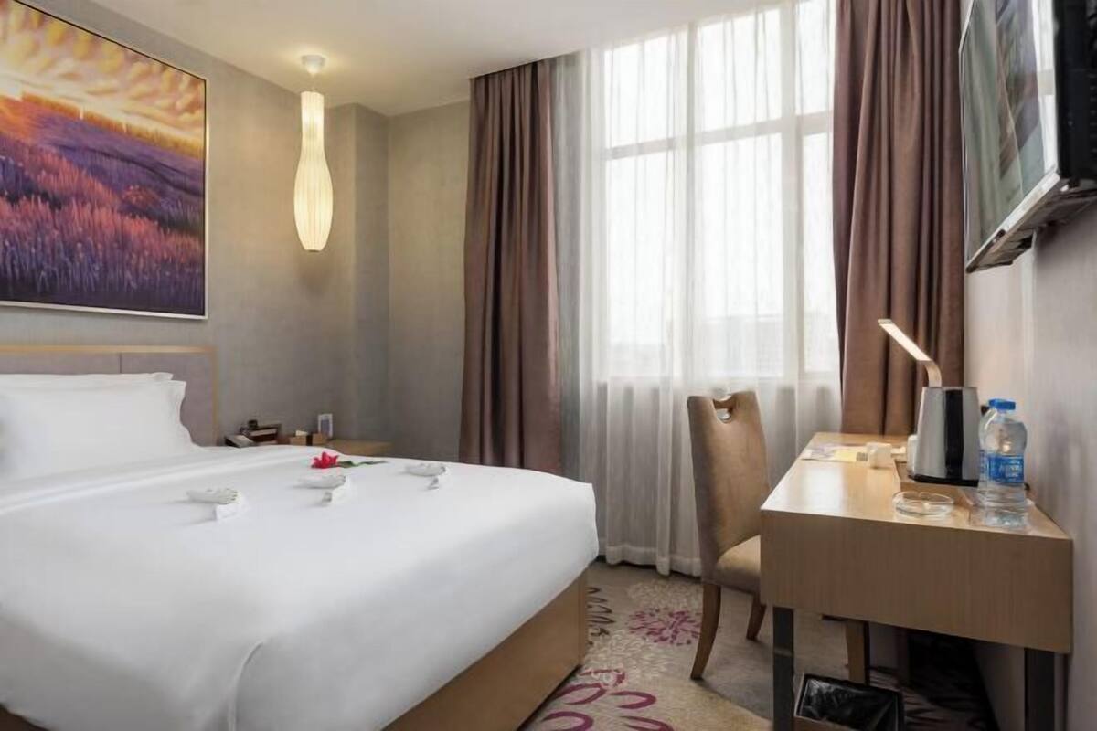 Quarto de hotel com cama, escrivaninha, cadeira, televisão e janela com cortinas.