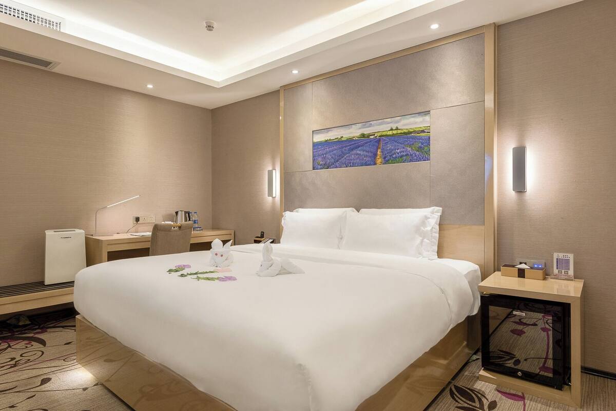 Quarto de hotel com uma cama grande, uma escrivaninha, uma cadeira, uma mesinha de cabeceira, um abajur e um quadro na parede.