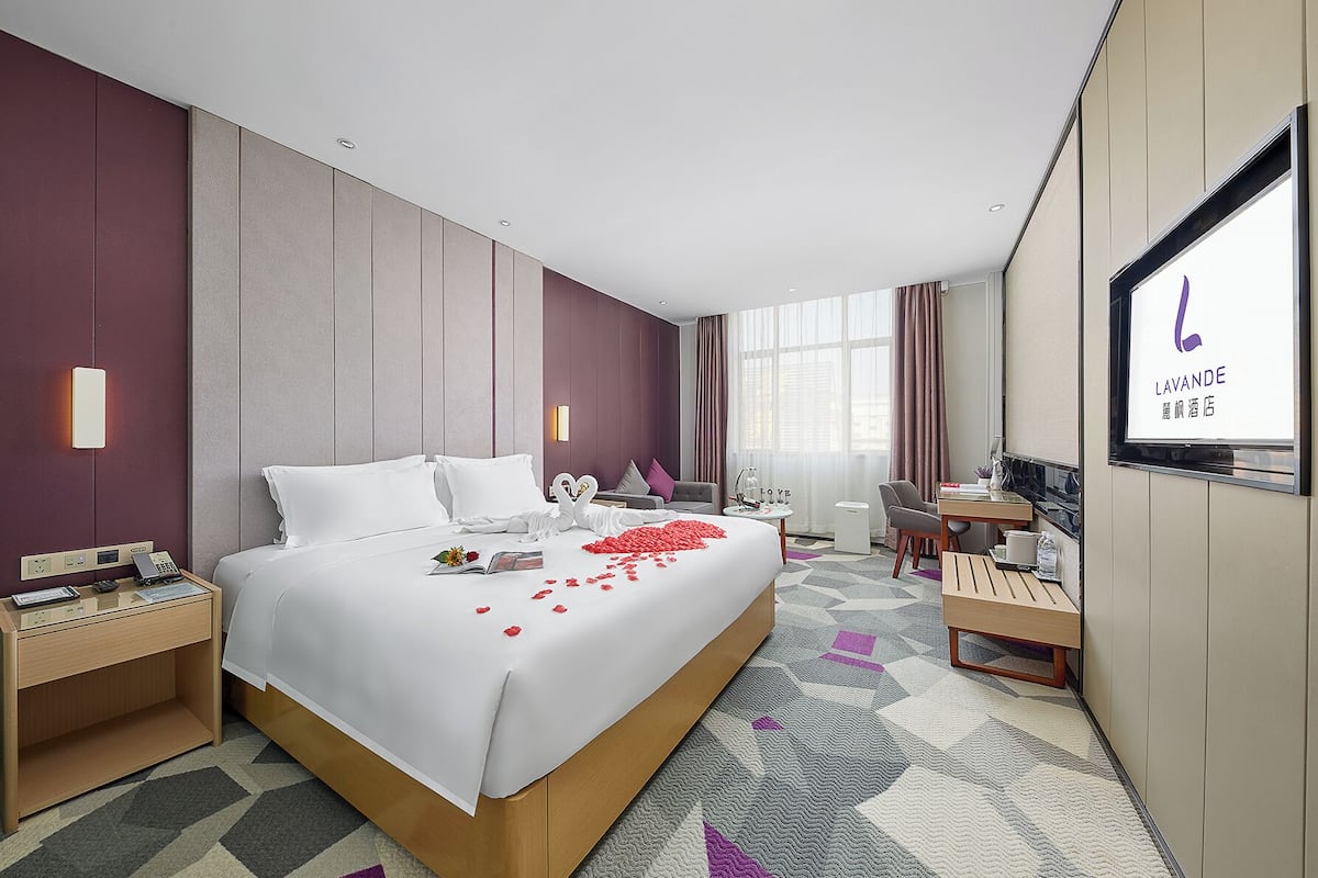 Quarto de hotel com uma cama grande, uma escrivaninha, uma cadeira, uma televisão e uma janela com cortinas.