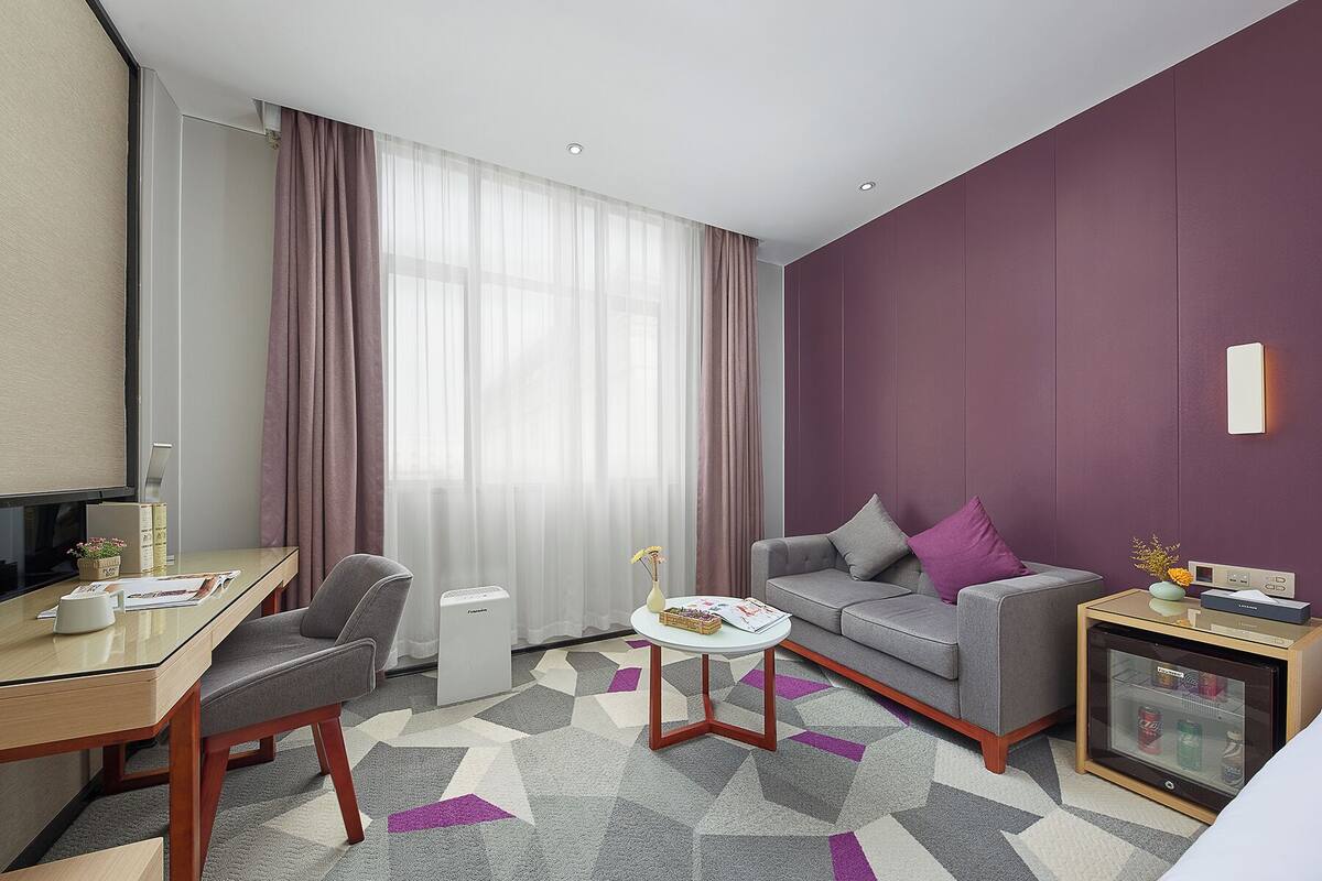 Quarto de hotel moderno com sofá cinzento, mesa redonda pequena, televisão e minibar.