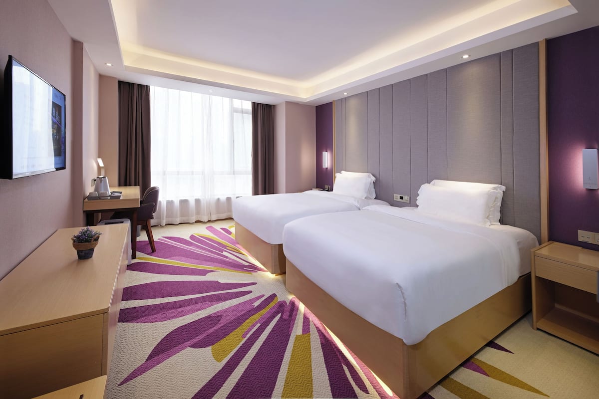 Quarto de hotel com duas camas, uma escrivaninha, uma televisão e uma janela grande com cortinas.
