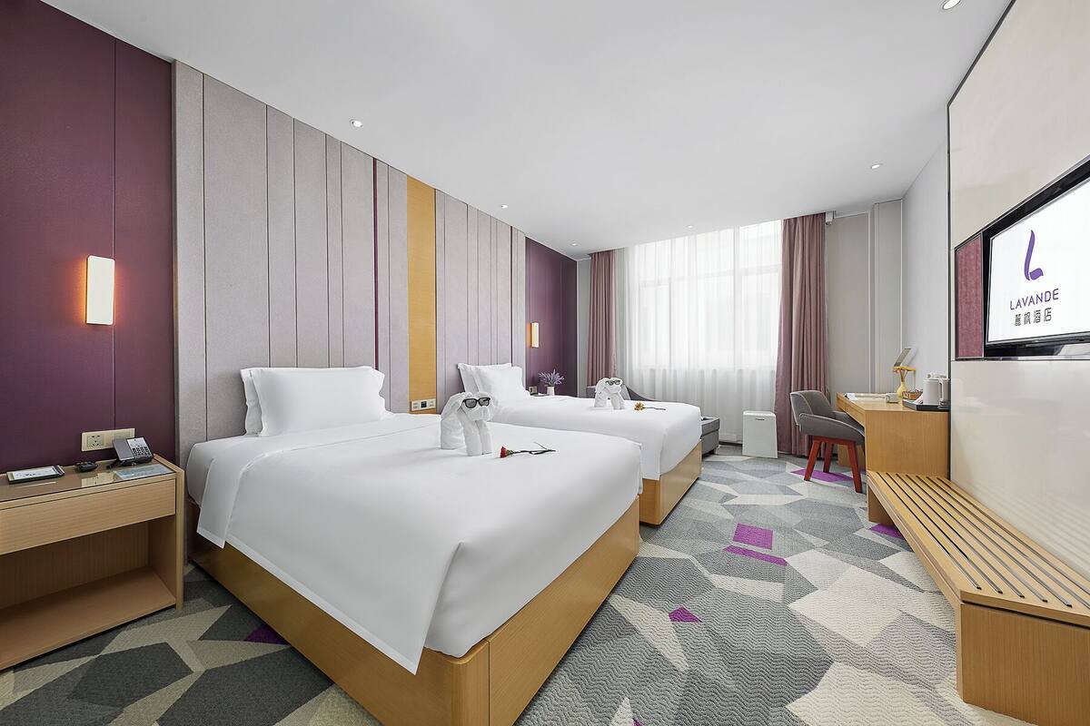 Quarto de hotel com duas camas, uma escrivaninha, uma cadeira, uma televisão e uma mesinha.