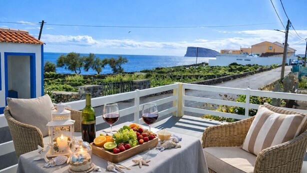 Uma varanda com móveis de vime, uma mesa posta com frutas, copos de vinho e uma vela, com vista para a costa.
