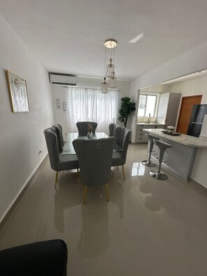 Een moderne keuken met een centraal eiland, een eethoek met een glazen tafel en grijze stoelen, en een groot raam met gordijnen.