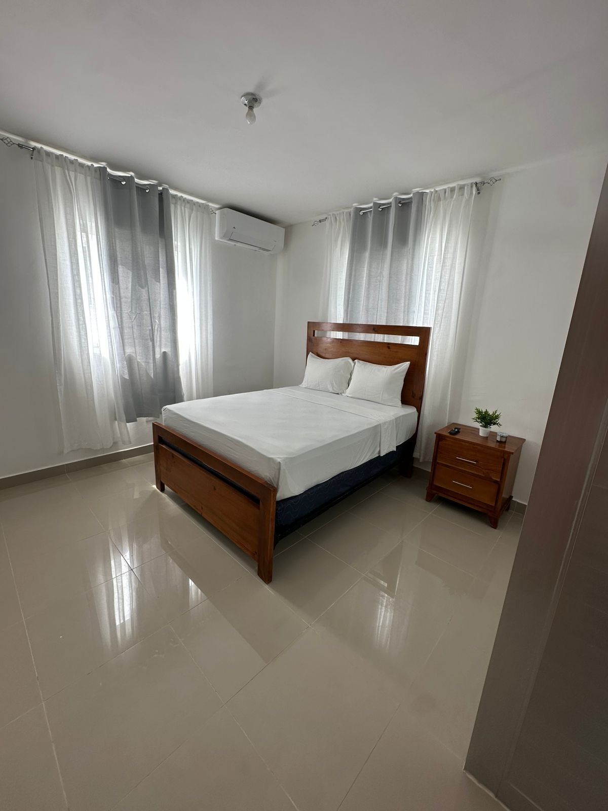 Une chambre avec un lit en bois, des rideaux blancs, une petite table de chevet avec une plante, et un appareil de climatisation fixé au mur.