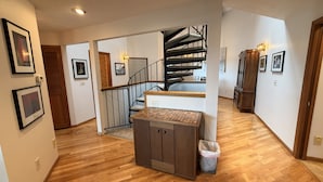 Un pasillo espacioso con piso de madera, una escalera con barandilla negra y un mueble pequeño con un basurero.