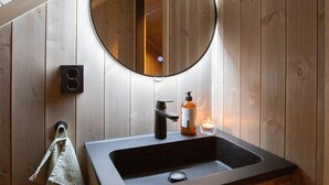 Een badkamer met een houten wand, een ronde spiegel, een zwarte wastafel, een zeepdispenser en een handdoek aan een haak.