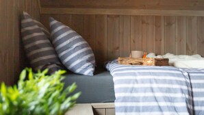 Een gezellige kamer met een houten plafond, een bed met gestreepte beddengoed, twee kussens, een rieten mand met een kop en schotel, en een zachte, witte deken.