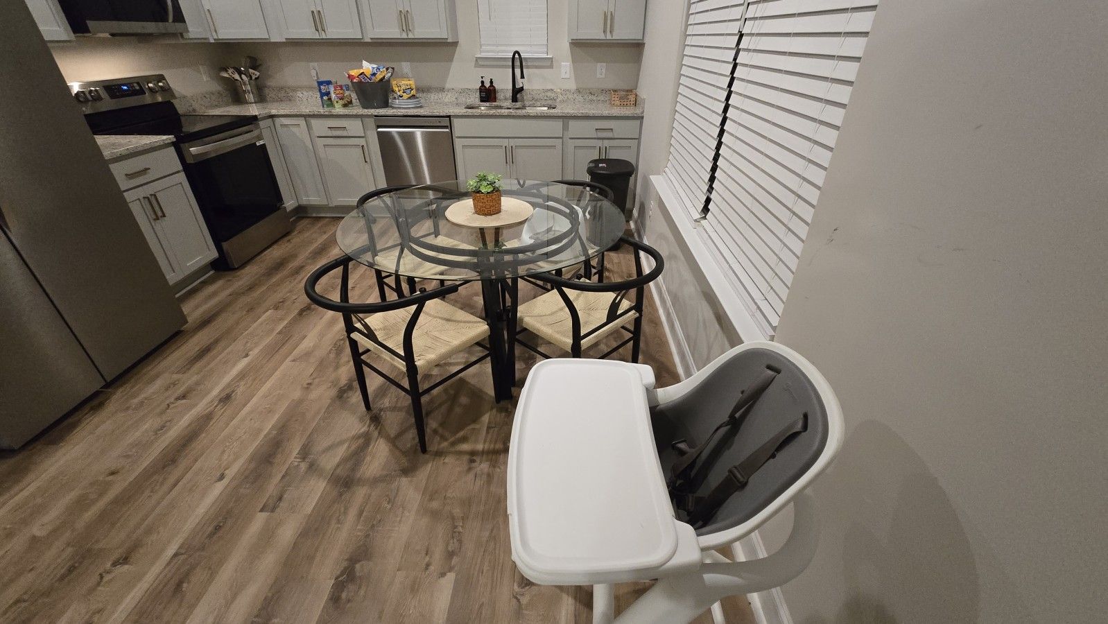 A compact kitchen with a dining area, featuring a round glass table, chairs, and a small white table with a black tray.
