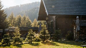 Un bâtiment rustique en bois, doté d’un toit en pente, entouré d’une zone boisée avec de petits arbres et, au loin, d’une montagne.