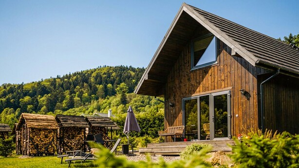 Une cabane en bois avec un toit en pente, une porte d’entrée en verre et une terrasse dotée de sièges extérieurs.