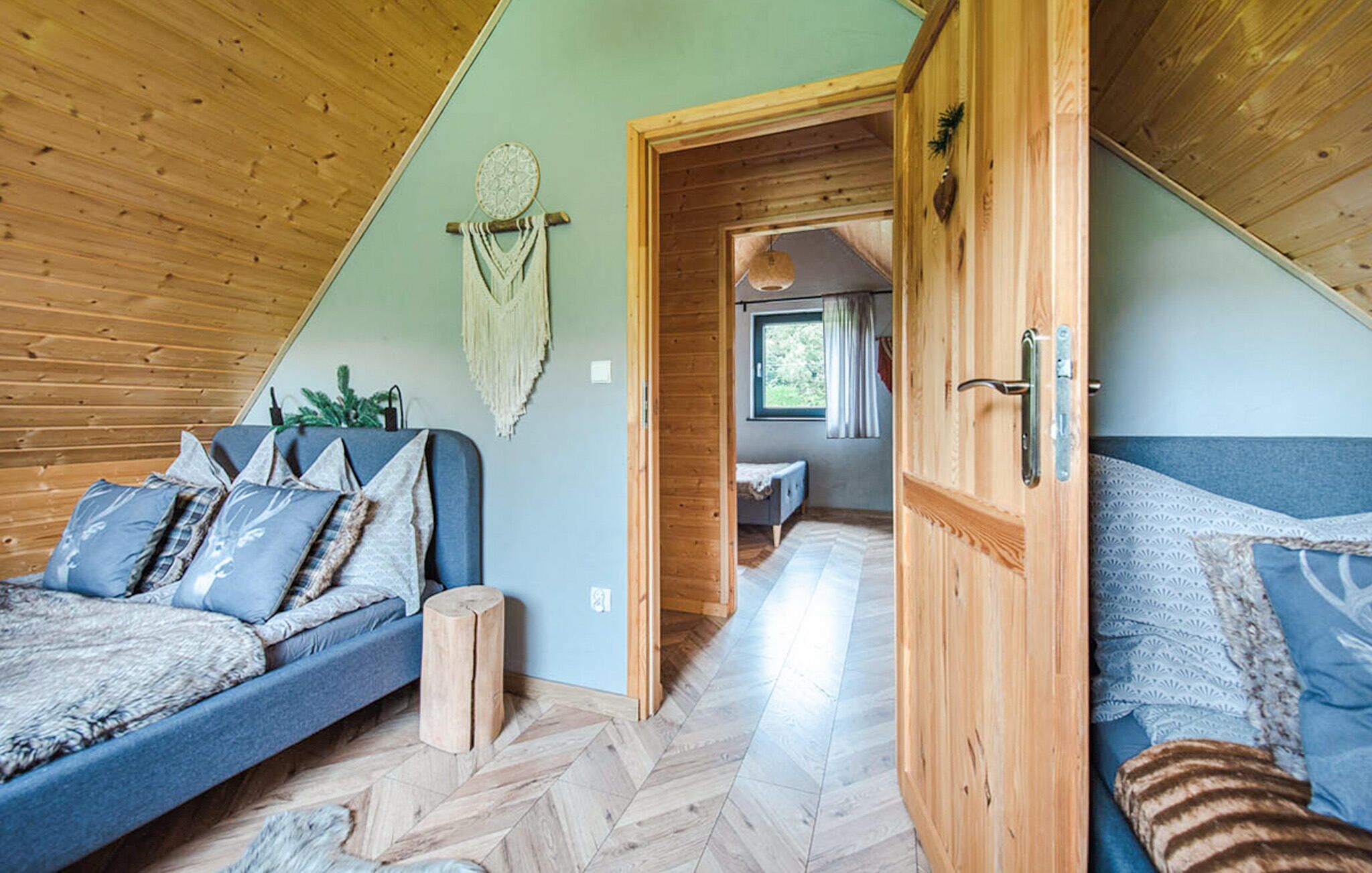 Een knus slaapkamer in loftstijl met een houten plafond, een groot bed met beddengoed met een patroon, een klein zithoekje en zicht door een deuropening naar een andere kamer.